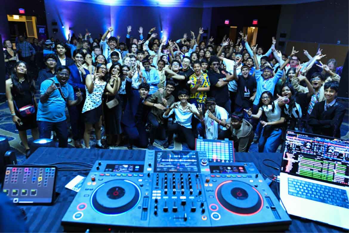 Large crowd posing behind a DJ booth with turntables and colorful stage lights in the background.