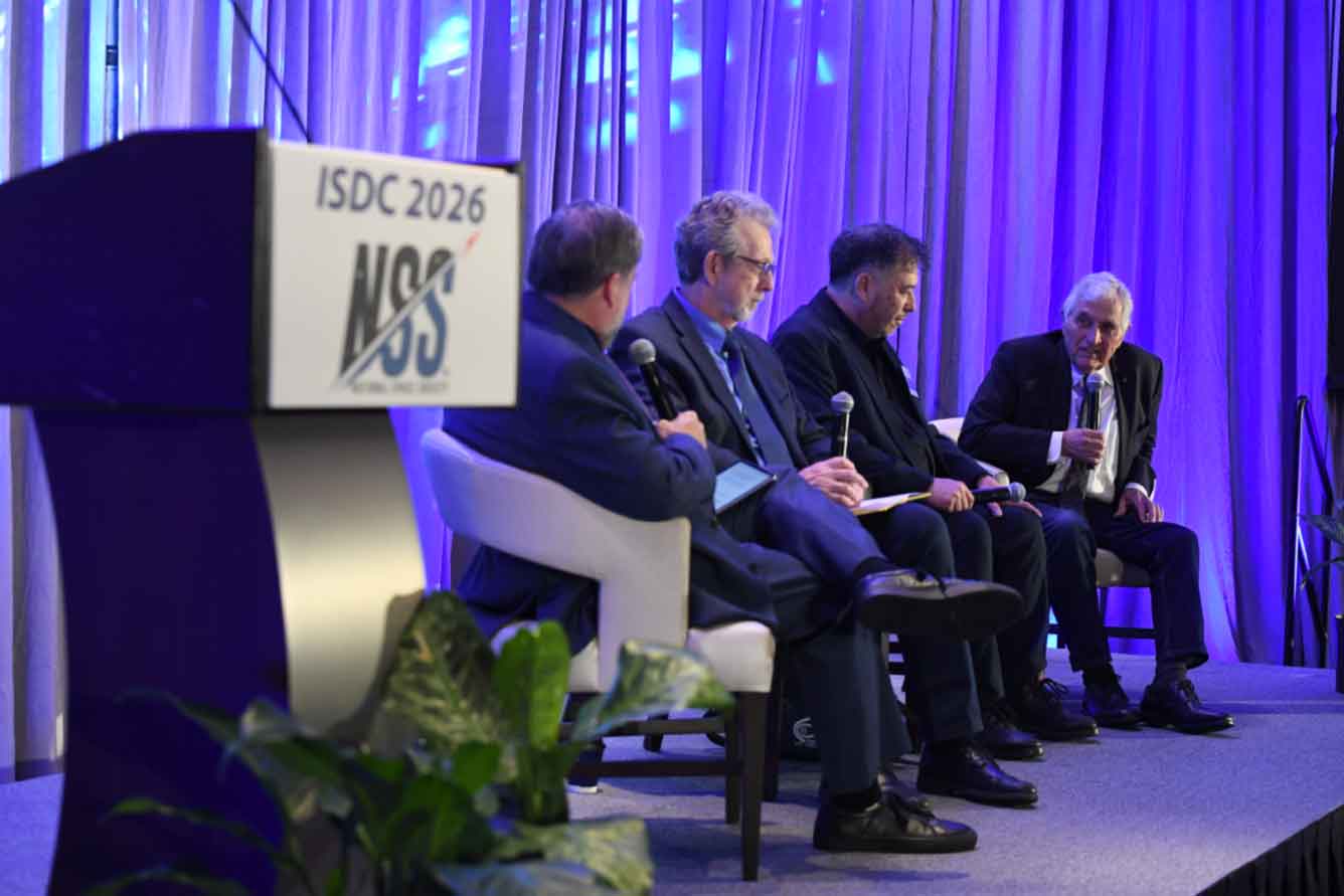 Panel of five men in suits seated on a stage with handheld microphones during a conference (ISDC 2026), blue-purple backdrop curtains behind them.