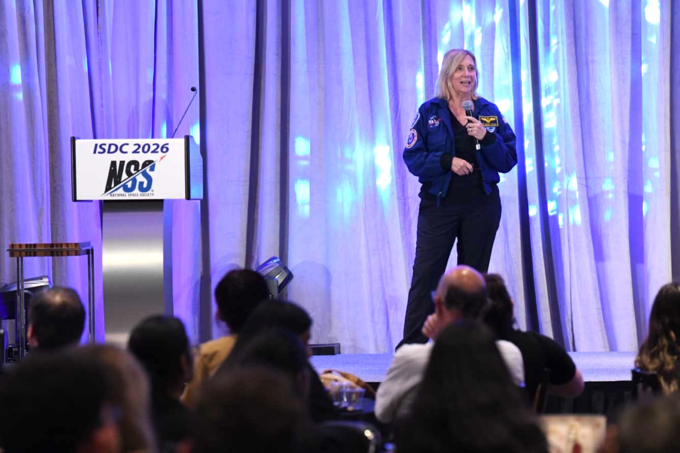 Woman in a blue flight jacket speaks into a microphone on a stage, addressing an audience at the ISDC 2026 event with a podium nearby.