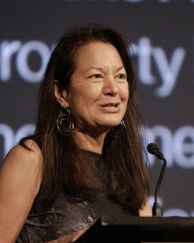Woman speaking at a podium on stage, microphone in front, wearing a dark sleeveless top and large hoop earrings.
