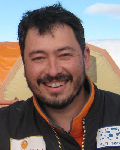 Smiling man with dark hair and beard wearing an orange-labeled jacket, outdoors at a campsite with tents in the background