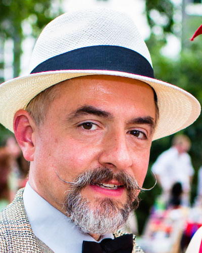 Samuel Coniglio | ISDC 2026 Man wearing a white straw hat with a dark band, gray moustache and beard, bow tie, outdoors at a social gathering.