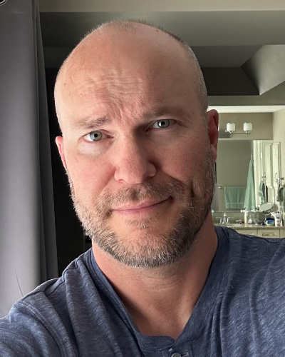 Close-up portrait of a smiling bald man with light stubble and blue eyes, wearing a blue shirt, indoors.