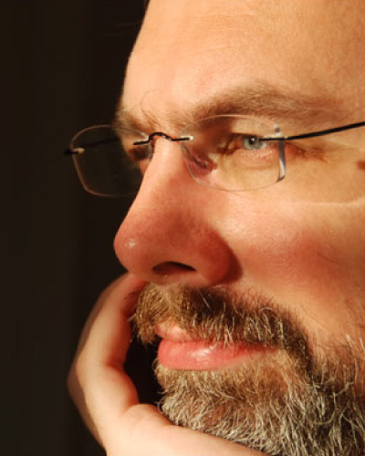 Teviet Creighton | ISDC 2026 Close-up of a bearded man wearing glasses, resting his chin on his hand and looking thoughtful.