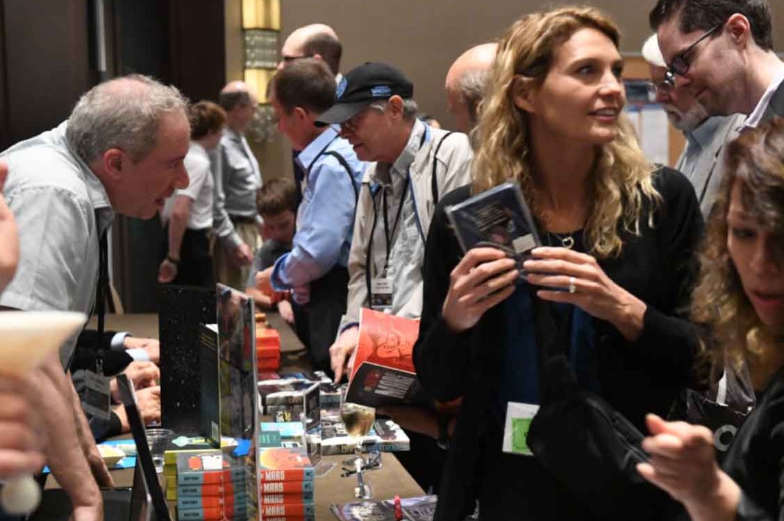 Blonde woman in a black jacket holding a brochure at a crowded expo booth with books and gadgets nearby