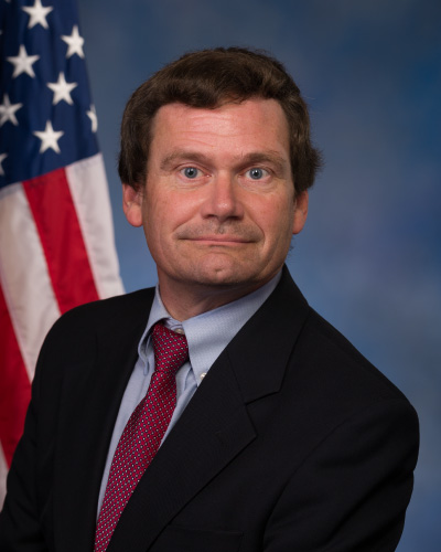 Professional portrait of a man in a dark suit and red tie with an American flag in the background.