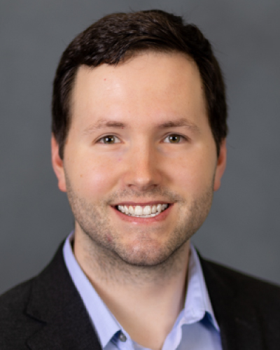 Professional headshot of a smiling man in a dark blazer and light blue shirt against a gray studio background