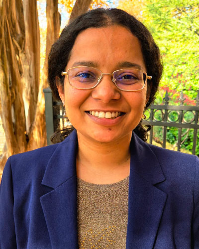 Smiling woman with short dark hair and glasses, wearing a navy blazer outdoors by a fence.