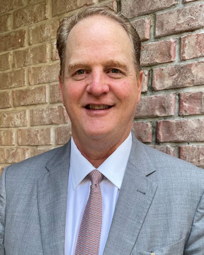 Head-and-shoulders portrait of a smiling man in a light gray suit and pink tie, standing in front of a brick wall/ backdrop of bricks.