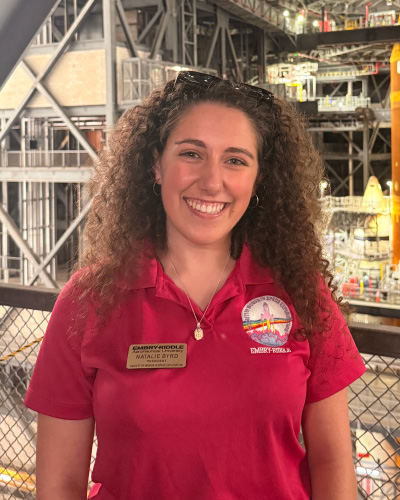 Smiling woman with curly hair in a red polo shirt, wearing a name badge and logo, sunglasses on her head, standing in a large industrial facility.