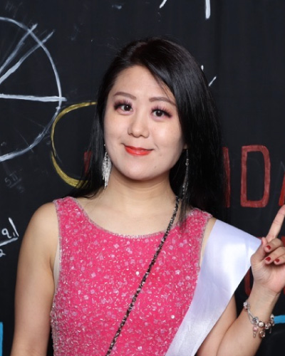 Woman in a pink sequined dress with a white sash smiles at the camera, making a peace sign; chalkboard backdrop behind her.