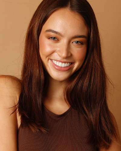 Smiling young woman with long brown hair wearing a sleeveless brown top, against a warm peach background.