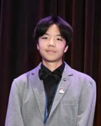 Young man in a light gray blazer and black bow tie stands on a stage with a dark curtain backdrop, wearing a name badge or pin on his lapel and a lanyard, smiling slightly.