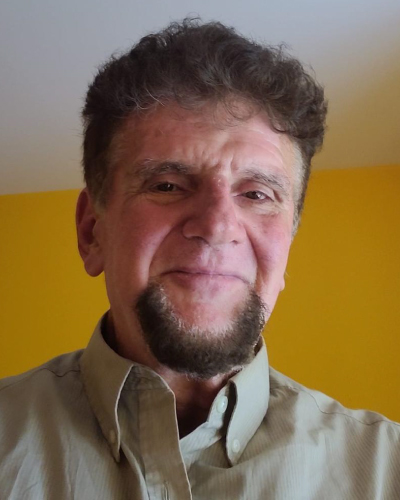 Smiling man with short curly hair and a goatee wearing a light beige shirt, standing in front of a bright yellow wall.