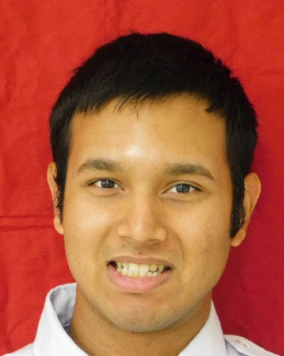 Close-up portrait of a man with short black hair, smiling at the camera, wearing a light collared shirt against a red backdrop.