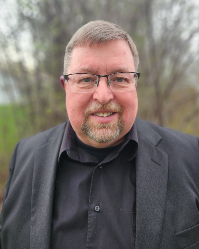 Smiling man with glasses in a dark blazer and black shirt, standing outdoors.