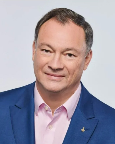Smiling man wearing a pink shirt and blue blazer against a light background.|Head-and-shoulders portrait in a blue suit and pink shirt, smiling.