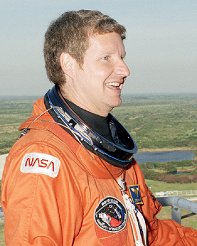 Profile view of a smiling astronaut in an orange NASA spacesuit with patches, helmet resting around the neck, outdoors with a lake and greenery in the background