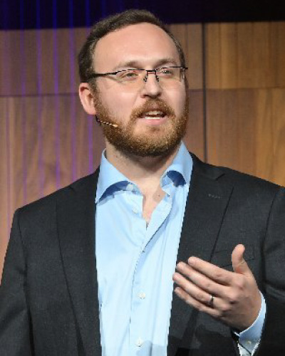 Bearded man in glasses giving a presentation on stage, wearing a light blue shirt and dark blazer, gesturing with his right hand.