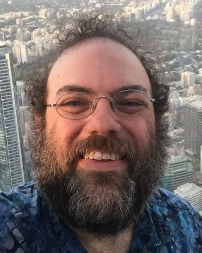 Close-up selfie of a man with glasses and a beard smiling, with a city skyline visible in the background.