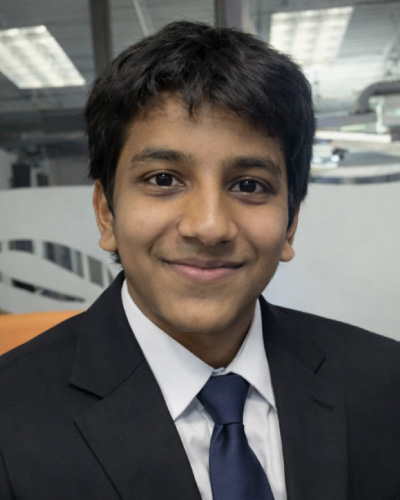 Young man in a dark suit and blue tie, smiling at the camera in an office setting.