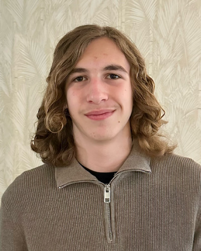 Portrait of a young man with shoulder-length wavy hair, wearing a tan zip-up sweater over a black shirt, standing against a light patterned wall.