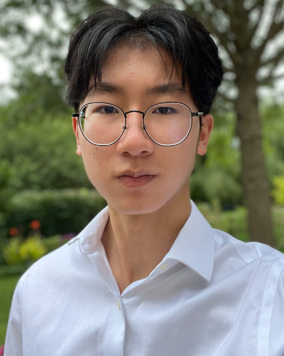 Portrait of a young man with round glasses and a white button-down shirt outdoors, neutral expression, green park in the background