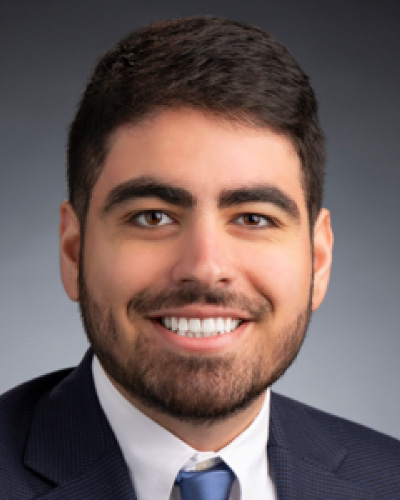 Head-and-shoulders portrait of a smiling man in a navy suit and blue tie against a gray gradient background.