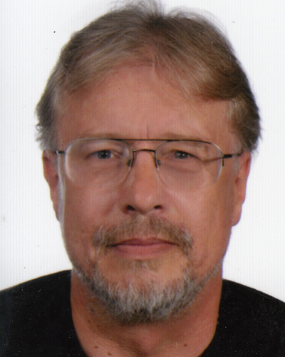 Close-up portrait of a middle-aged man with gray beard and metal-framed glasses, facing the camera.