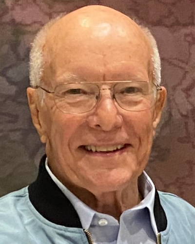 Smiling older man with glasses, bald head, wearing a light blue shirt indoors.