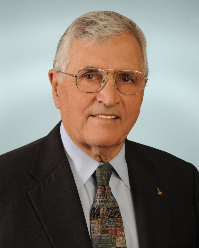 Professional portrait of a smiling older man with gray hair and glasses, wearing a dark suit, light blue shirt, and patterned tie.