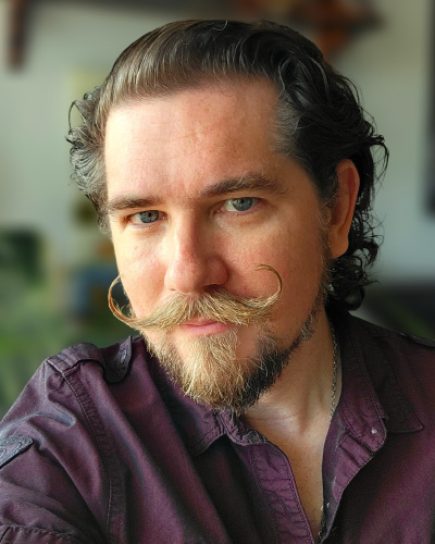 Portrait of a man with a curled handlebar mustache and goatee, wearing a burgundy shirt and looking at the camera.