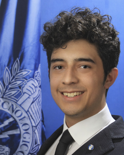 Young man in a suit and tie smiling in front of a blue backdrop with a school emblem.