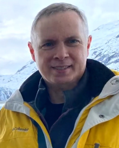 Smiling man in a bright yellow winter jacket outdoors in a snowy mountain landscape.
