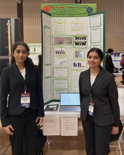 Two young women in blazers stand beside a science-fair poster board titled Red Planet Green Future, with charts and a laptop on the table.