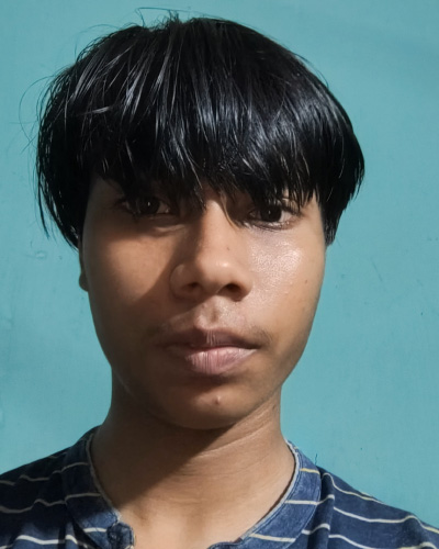 Portrait of a young person with short black hair wearing a striped blue shirt, standing in front of a teal wall and looking at the camera.