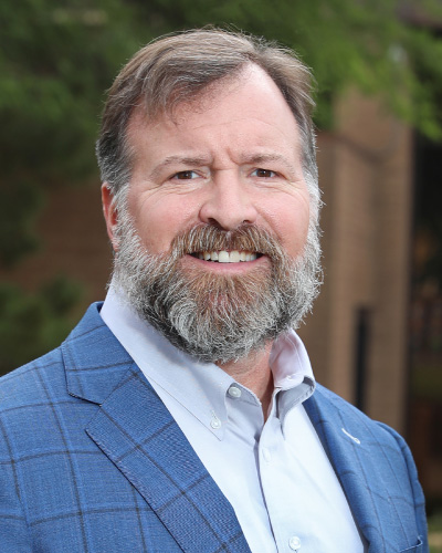 Portrait of a smiling man with a gray beard wearing a blue checkered blazer and light blue shirt, outdoors with greenery in the background