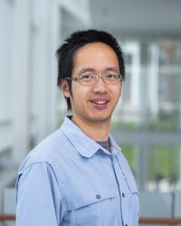 Smiling man wearing a light blue button-up shirt and glasses, standing in a modern building hallway.