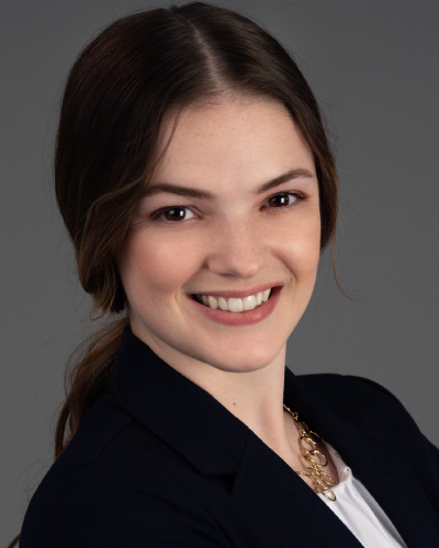 Portrait of a smiling woman with brown hair tied back, wearing a navy blazer and white top, against a gray backdrop.