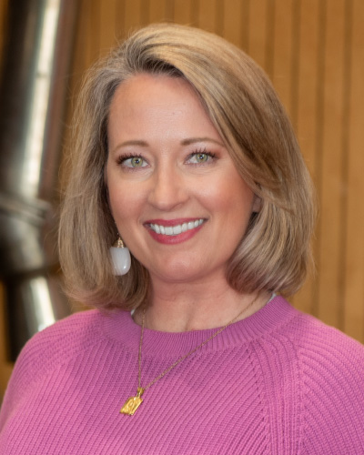 Amy McDowell | ISDC 2026 Portrait of a smiling woman with shoulder-length blonde hair, wearing a pink sweater and gold necklace indoors.