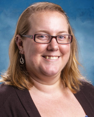 Angela Stickle | ISDC 2026 Portrait of a smiling woman with light brown hair, glasses, and teardrop earrings, wearing a dark cardigan against a blue backdrop
