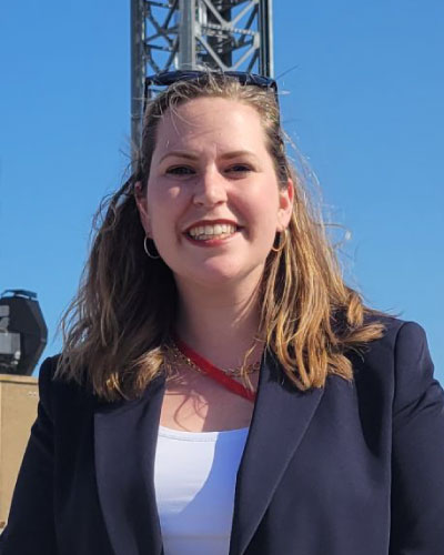 Smiling woman with shoulder-length hair in a navy blazer and white top, sunglasses on head, outdoors with a metal tower and clear blue sky behind her.