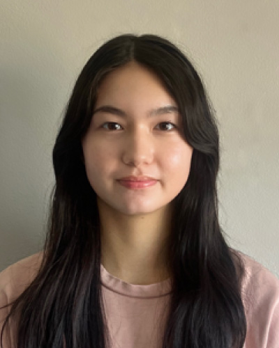 Portrait of a young Asian woman with long dark hair, wearing a light pink shirt, smiling softly against a neutral wall.