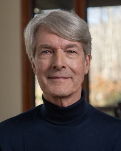 Portrait of an older man with silver hair in a black turtleneck, facing the camera with a neutral expression indoors.