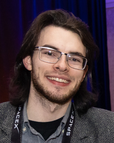 Smiling man with shoulder-length dark hair and glasses, wearing a blazer and a lanyard at an event.