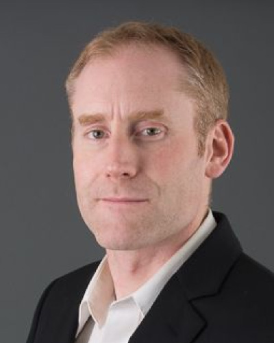 Professional headshot: man in a black blazer and white shirt against a gray background.