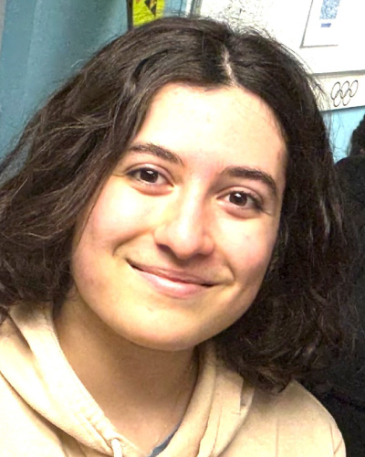 Young woman with wavy dark hair smiling at the camera, wearing a beige hoodie overseeing a casual indoor setting.