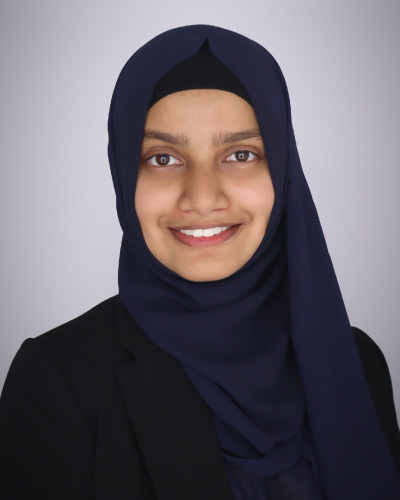 Sahda Haroon | ISDC 2026 Portrait of a smiling woman wearing a navy hijab and black blazer against a light gray background.