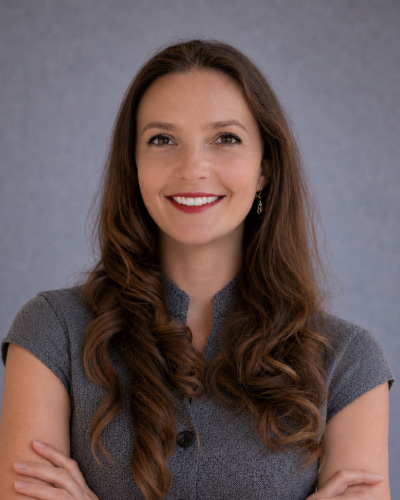 Irina Litchfield | ISDC 2026 Portrait of a smiling woman with long brown hair, wearing a gray short-sleeve top, arms crossed.