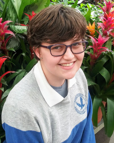 Jaclyn Wiley | ISDC 2026 Smiling person with short brown hair and glasses in a gray-blue polo, standing among red tropical flowers and green leaves.
